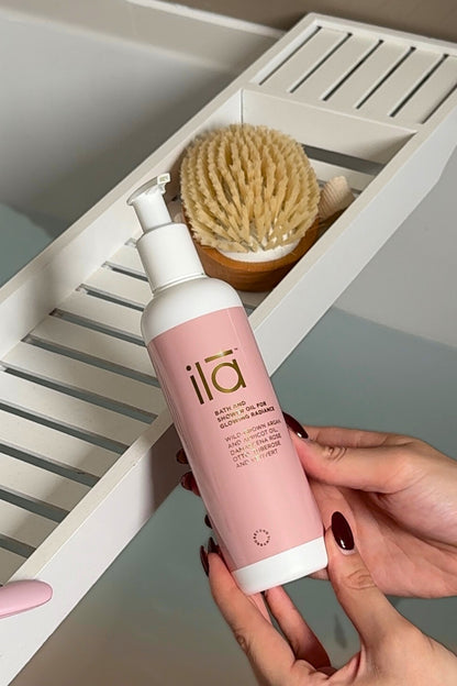 Person holding a pink and white bottle labeled 'ila' in front of a bathroom shelf with a brush.