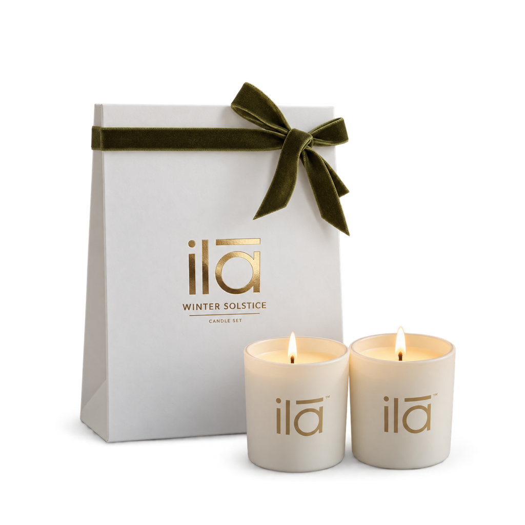Two cream-colored candles with gold 'ila' branding in front of a white gift bag tied with an olive green velvet ribbon. The brand 'ila' and 'WINTER SOLSTICE' are printed in gold on the bag.