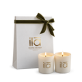 Two cream-colored candles with gold 'ila' branding in front of a white gift bag tied with an olive green velvet ribbon. The brand 'ila' and 'WINTER SOLSTICE' are printed in gold on the bag.