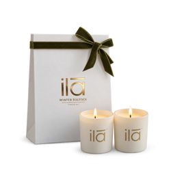 Two cream-colored candles with gold 'ila' branding in front of a white gift bag tied with an olive green velvet ribbon. The brand 'ila' and 'WINTER SOLSTICE' are printed in gold on the bag.